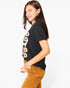 A woman wearing a black graphic t-shirt with a colorful print and brown corduroy pants, photographed against a solid white background.