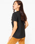 A woman shown from the back wearing a black short-sleeve t-shirt with a gold graphic print on the left side and brown corduroy pants, against a plain white background.