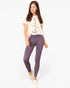 A woman modeling a light pink graphic t-shirt and high-waisted mauve-purple leggings with white slip-on sneakers, set against a plain white studio background.