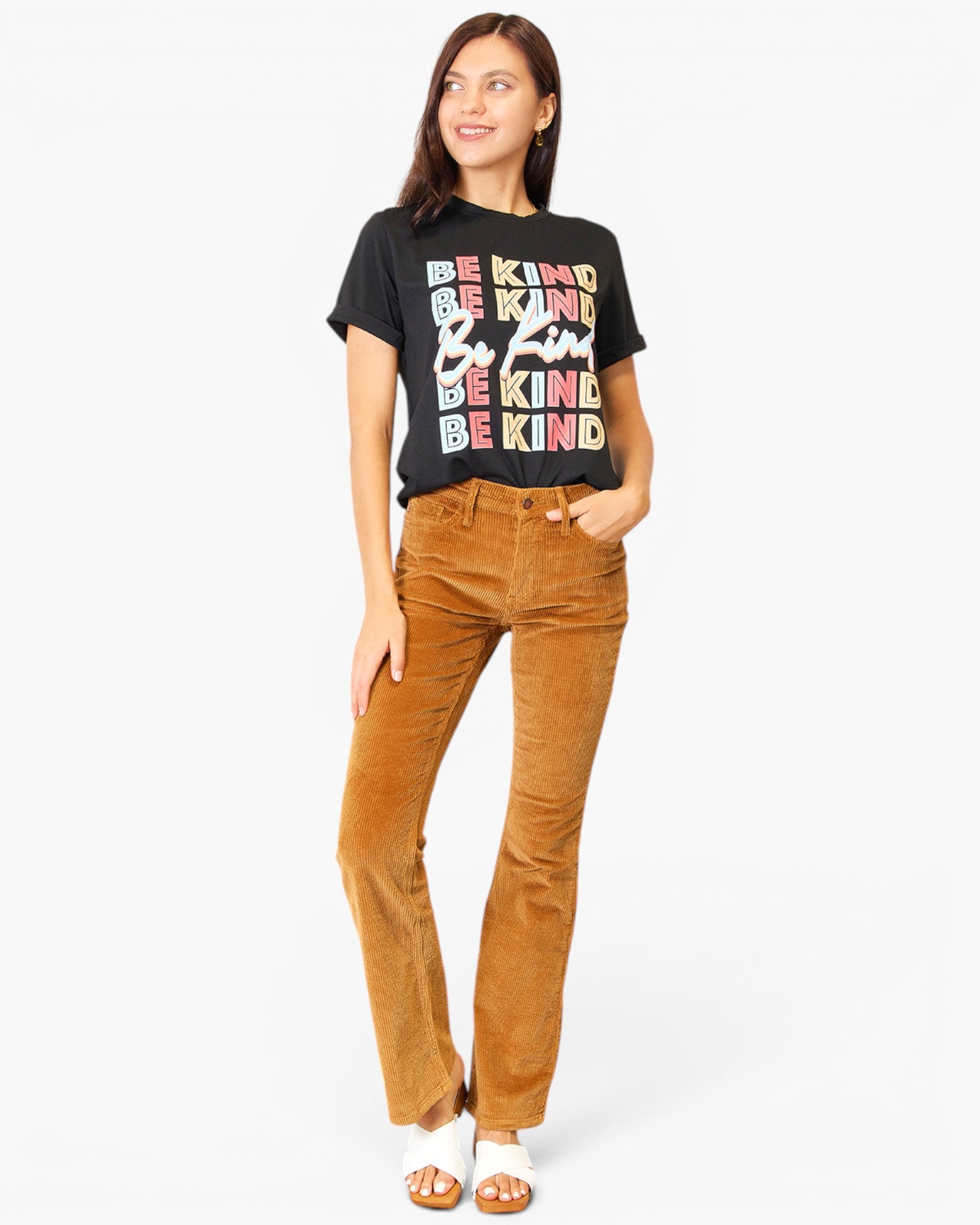 A woman modeling a black graphic t-shirt with stacked multicolor text reading 'BE KIND' and brown corduroy flare pants, paired with white slide sandals, against a plain white studio background.