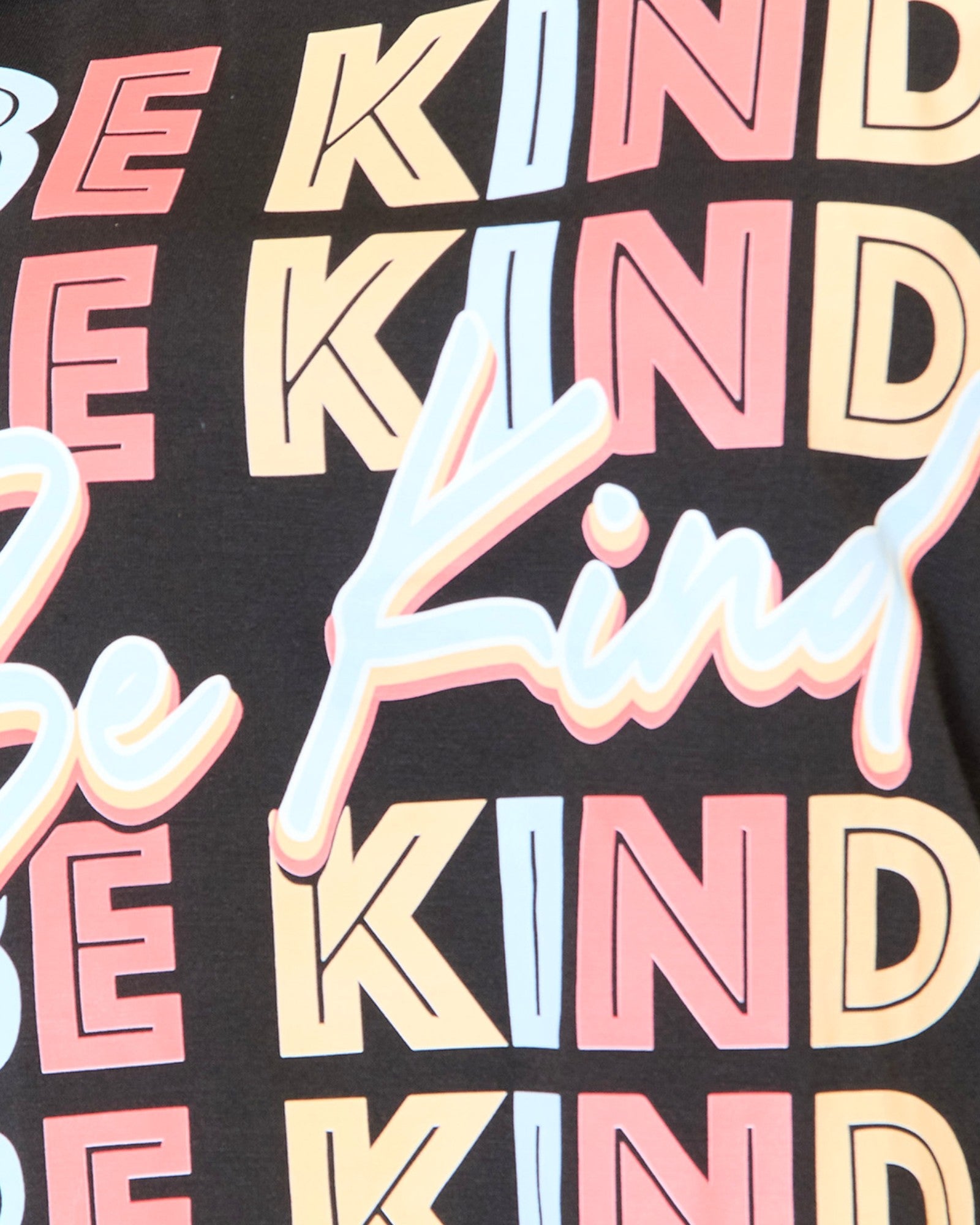 Close-up of a black graphic T-shirt featuring the word 'KIND' repeated in pastel pink, cream, and light blue, overlaid with a large white script word 'Be' outlined in pink.