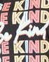 Close-up of a black graphic T-shirt featuring the word 'KIND' repeated in pastel pink, cream, and light blue, overlaid with a large white script word 'Be' outlined in pink.