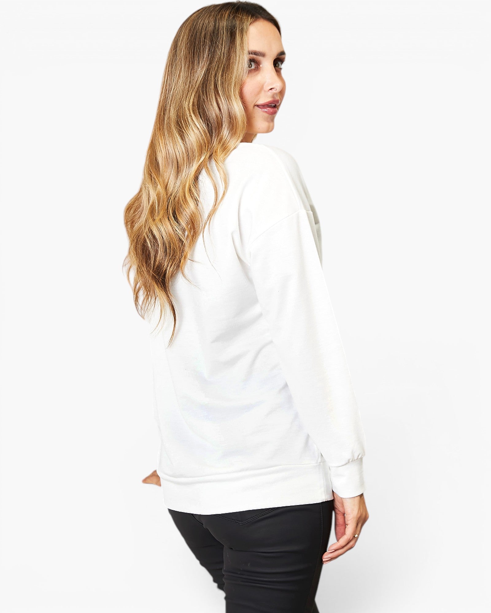 A woman with long wavy blonde hair is shown from the back wearing a white long-sleeve crewneck sweatshirt and black pants against a plain white studio background.