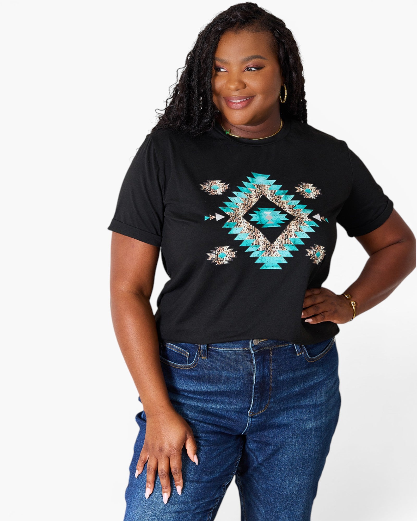 A woman modeling a black short-sleeve t-shirt with a colorful geometric diamond pattern in turquoise, beige, and brown on the front, paired with blue jeans against a plain white background.