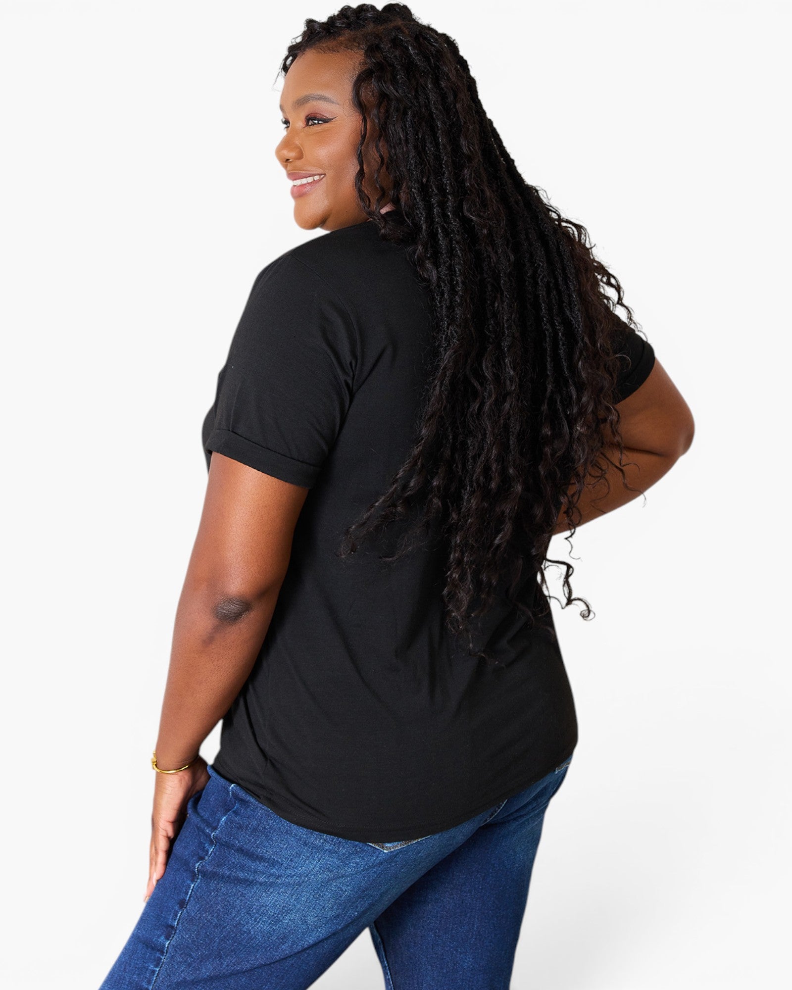 A woman with long, curly black hair is shown from a back-three-quarter angle wearing a black short-sleeve t-shirt and blue jeans against a plain white studio background.