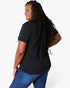 A woman with long, curly black hair is shown from a back-three-quarter angle wearing a black short-sleeve t-shirt and blue jeans against a plain white studio background.