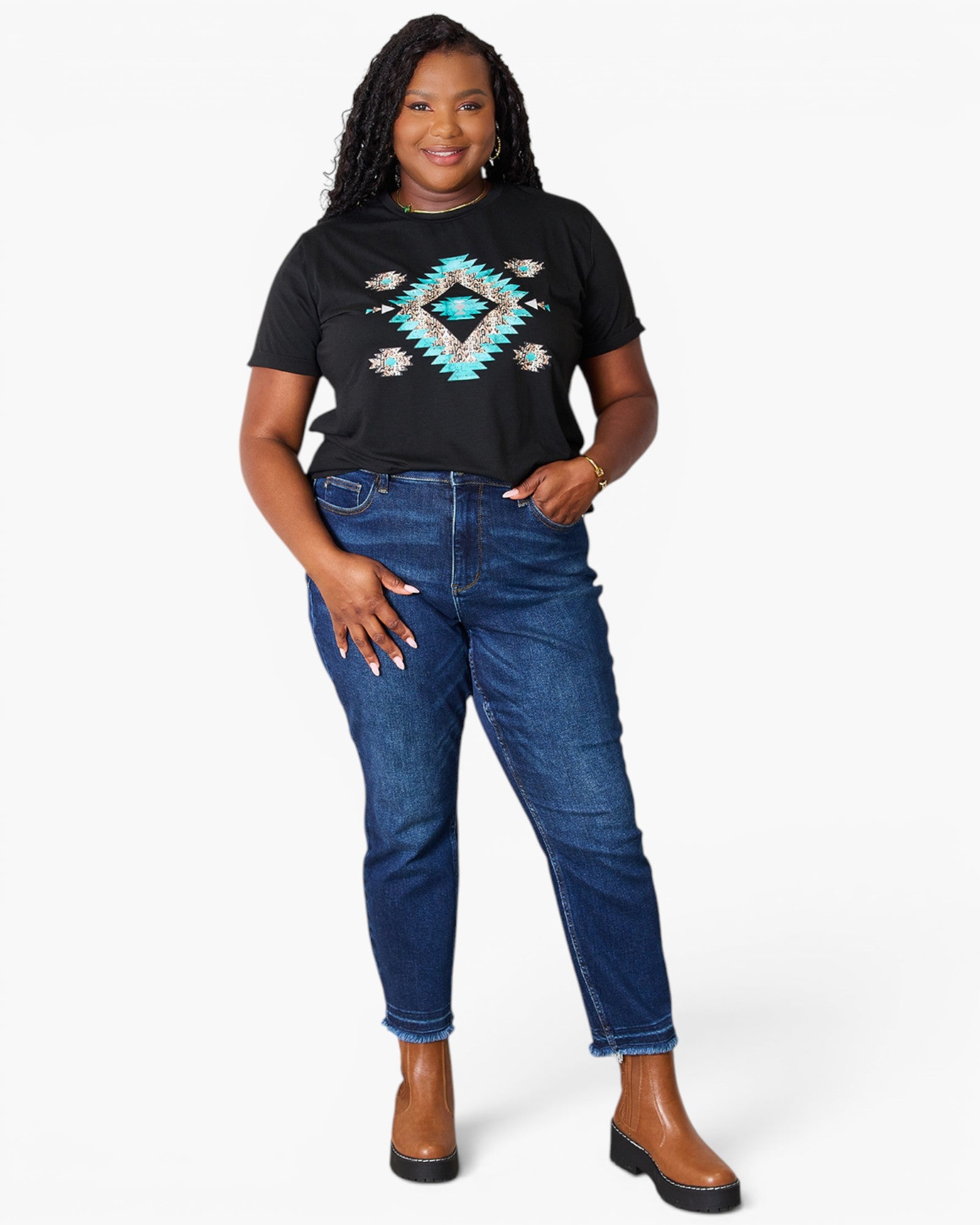 A woman modeling a black short-sleeve graphic t-shirt with a turquoise and white geometric diamond pattern, paired with dark blue jeans and brown ankle boots, against a light gray studio background.
