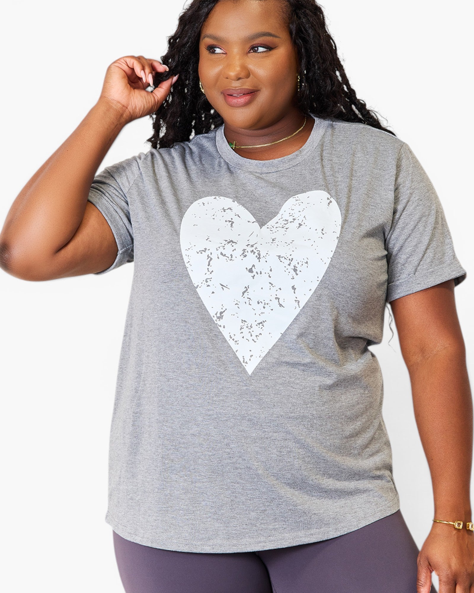 A woman wearing a heather gray short-sleeve t-shirt with a large distressed white heart graphic on the front, paired with purple leggings, against a plain white background.