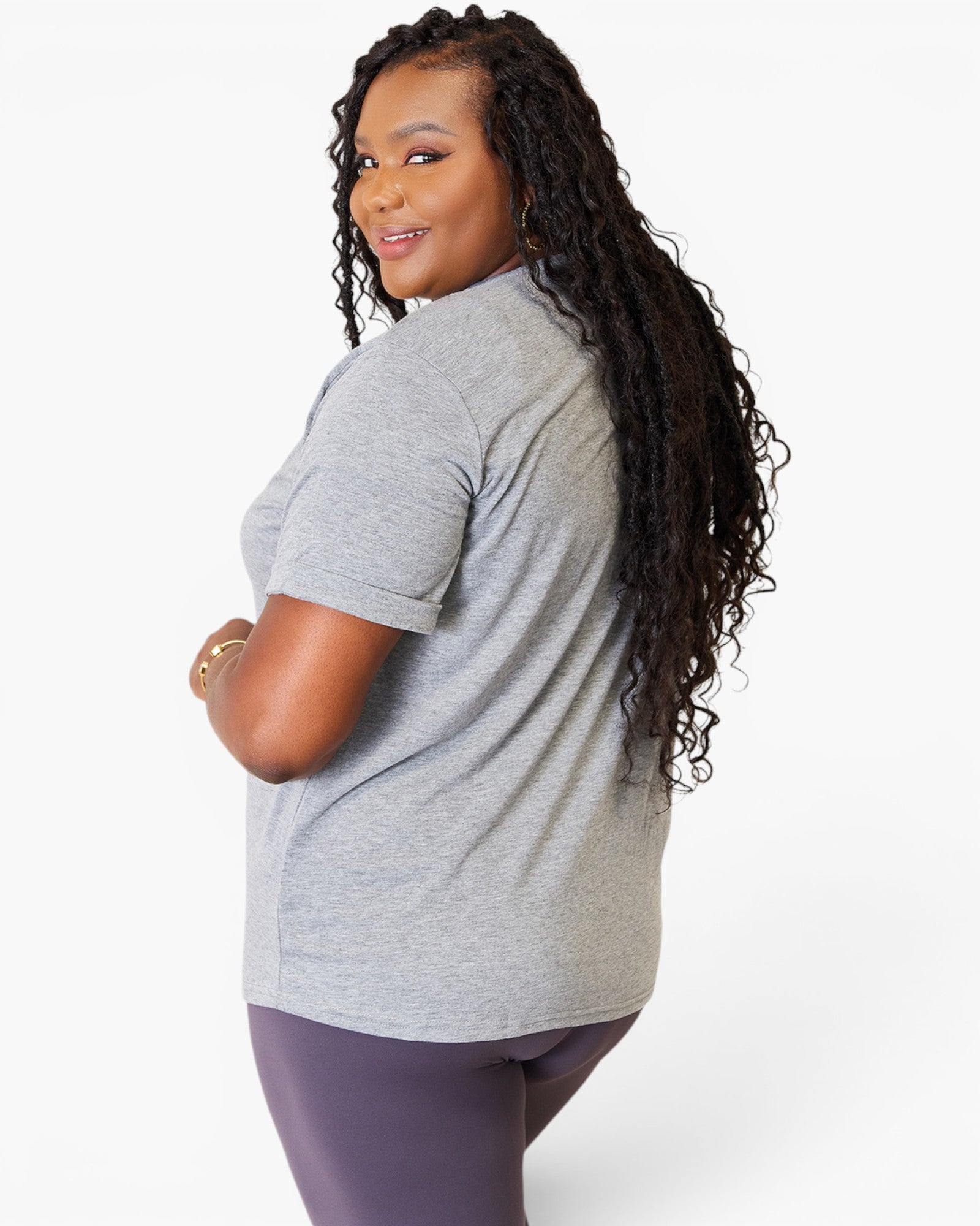 A woman with long, curly black hair is shown from a side-back angle wearing a light gray heather short-sleeve athletic t-shirt and dark purple leggings against a clean white studio background.