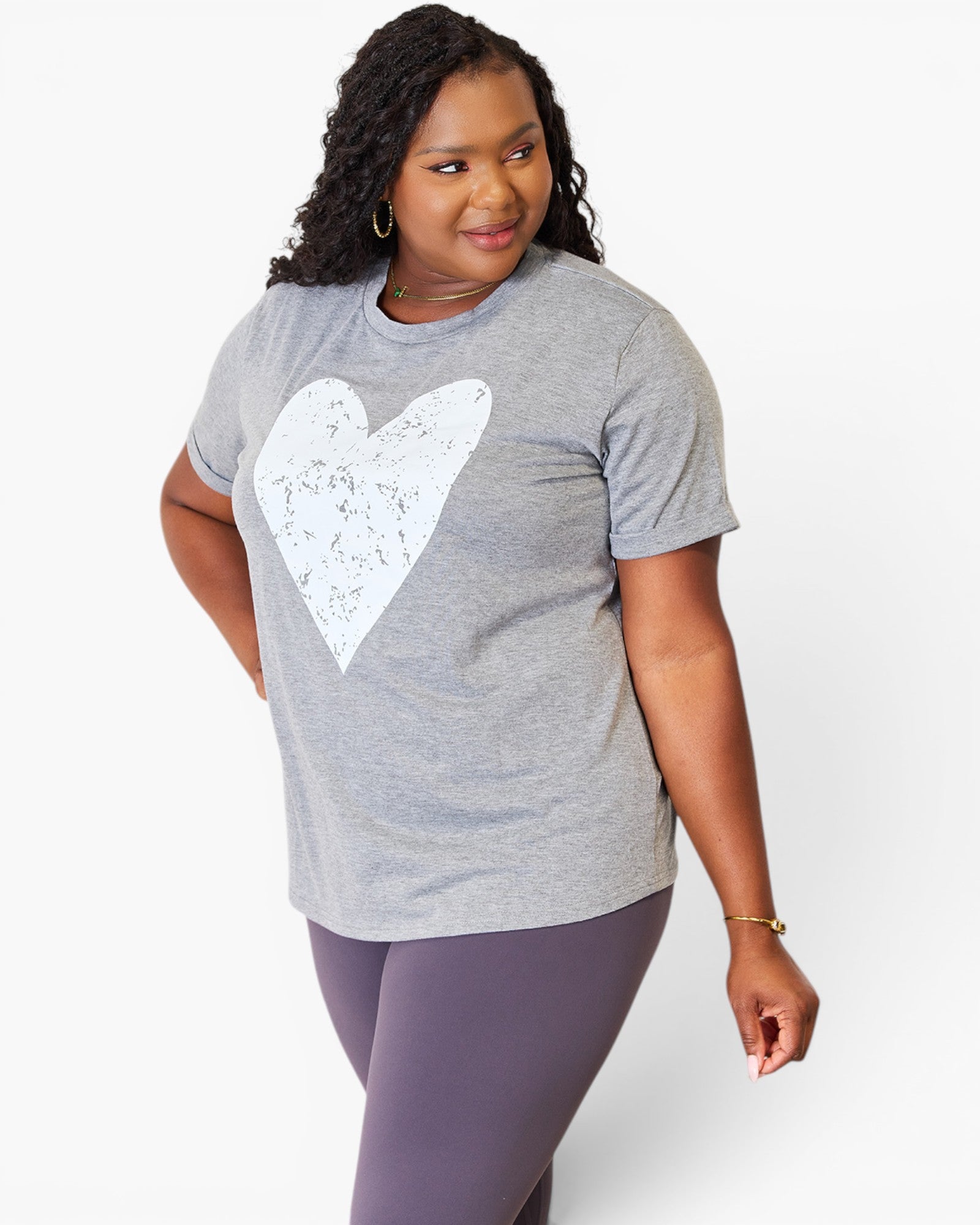 A woman modeling a heather gray short-sleeve t-shirt with a large white distressed heart graphic on the front, paired with purple leggings, against a light gray studio background.