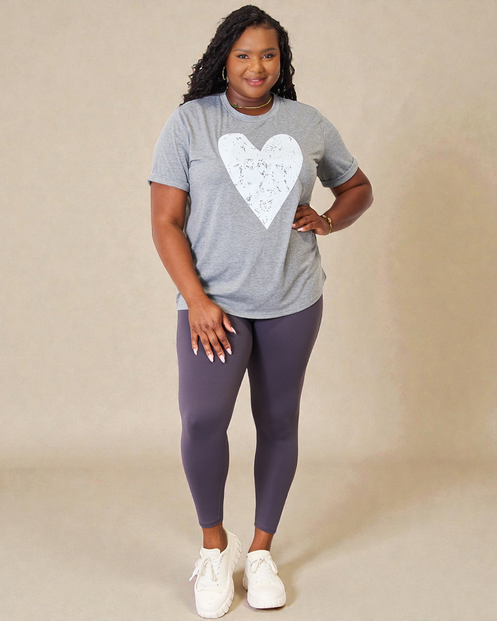 Woman wearing a gray t-shirt with a white heart design and purple leggings on a beige background