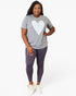 A woman modeling a heather gray t-shirt with a white heart graphic and dark purple leggings, paired with white sneakers, against a plain white studio background.