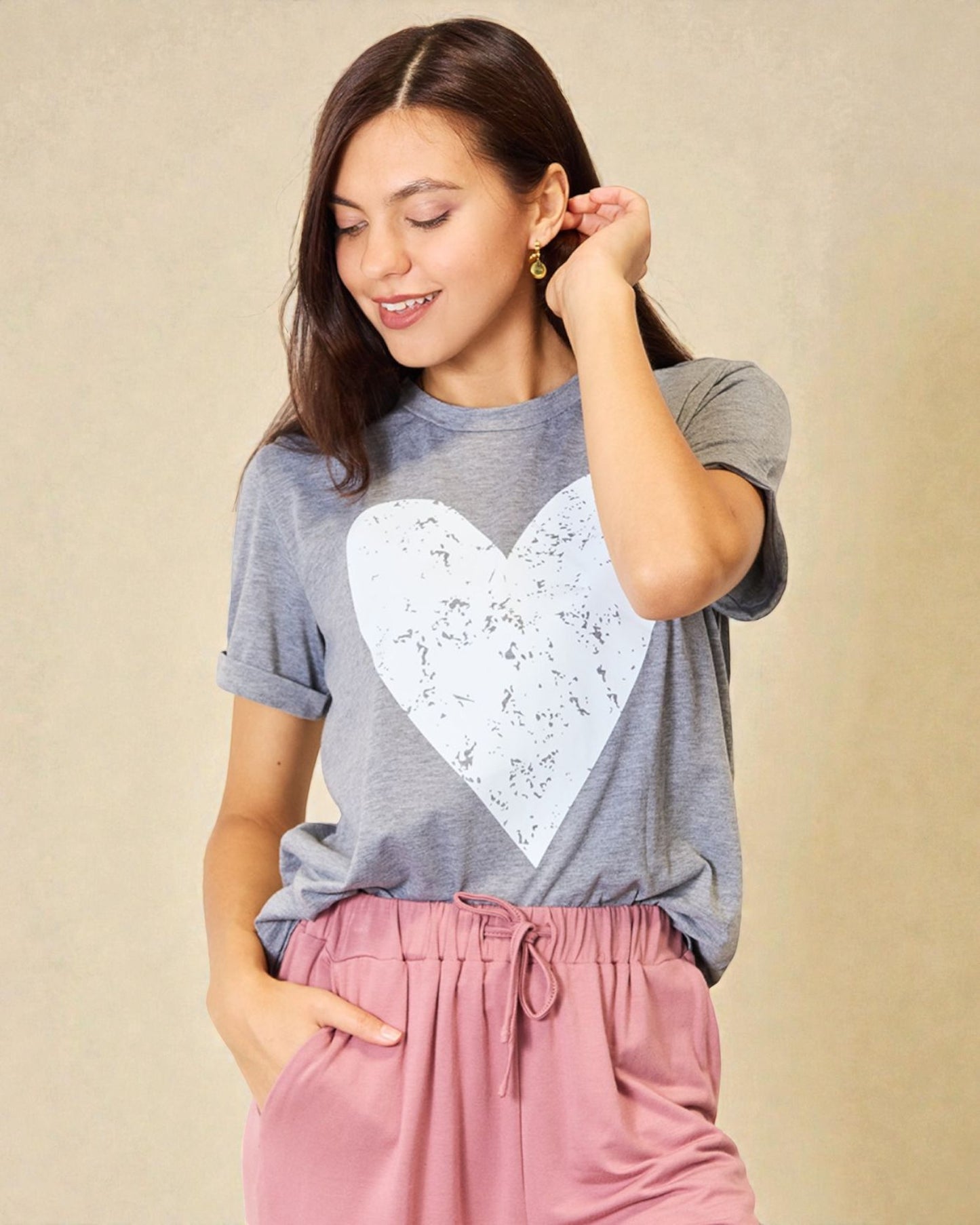 Woman wearing a gray t-shirt with a white heart design and pink pants against a beige background