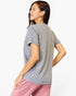 A woman wearing a heather gray short-sleeve t-shirt and pink pants, shown from a three-quarters back view against a plain white studio background.
