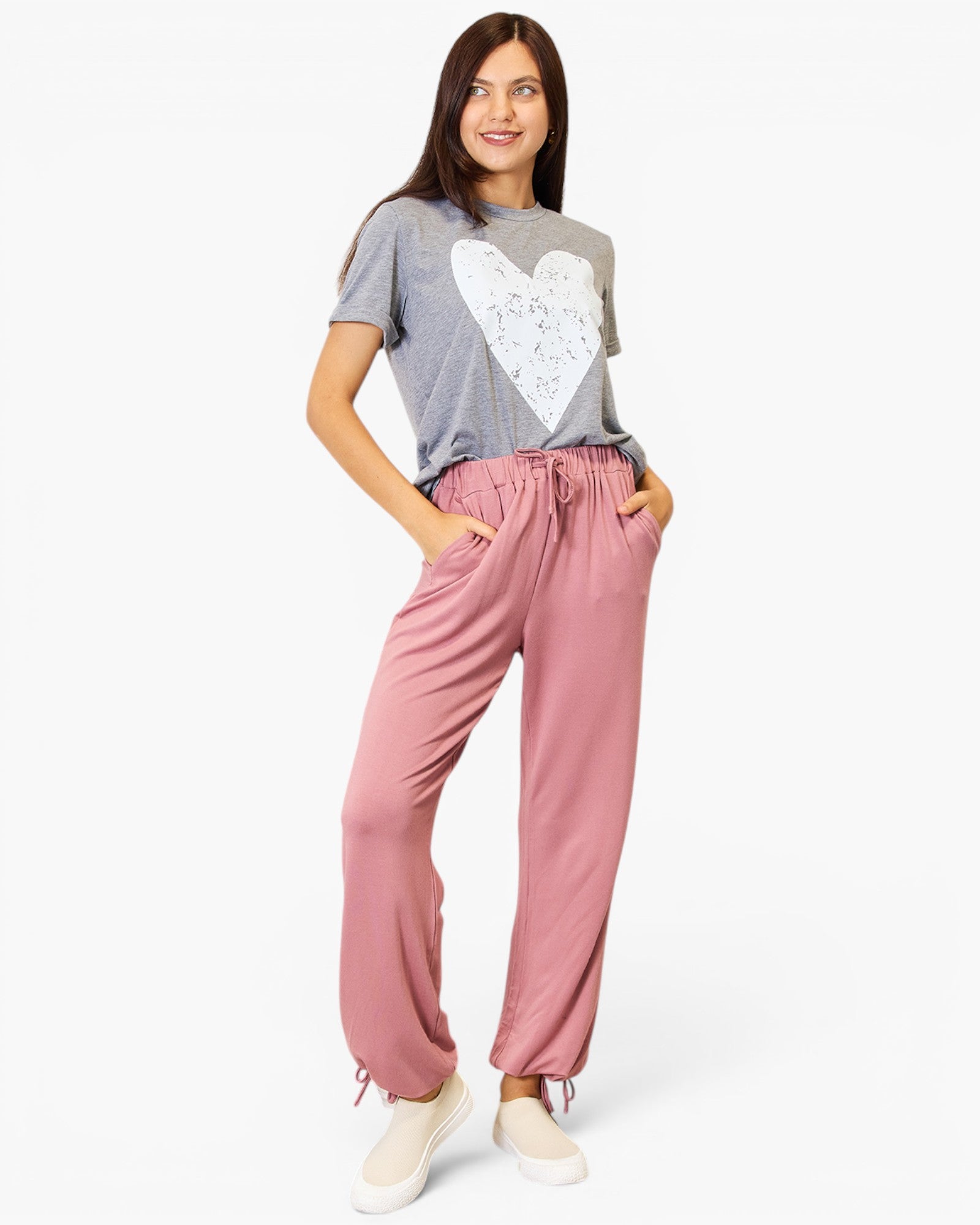 A woman modeling a gray t-shirt with a white heart graphic and dusty pink drawstring pants, paired with white sneakers, against a plain white background.