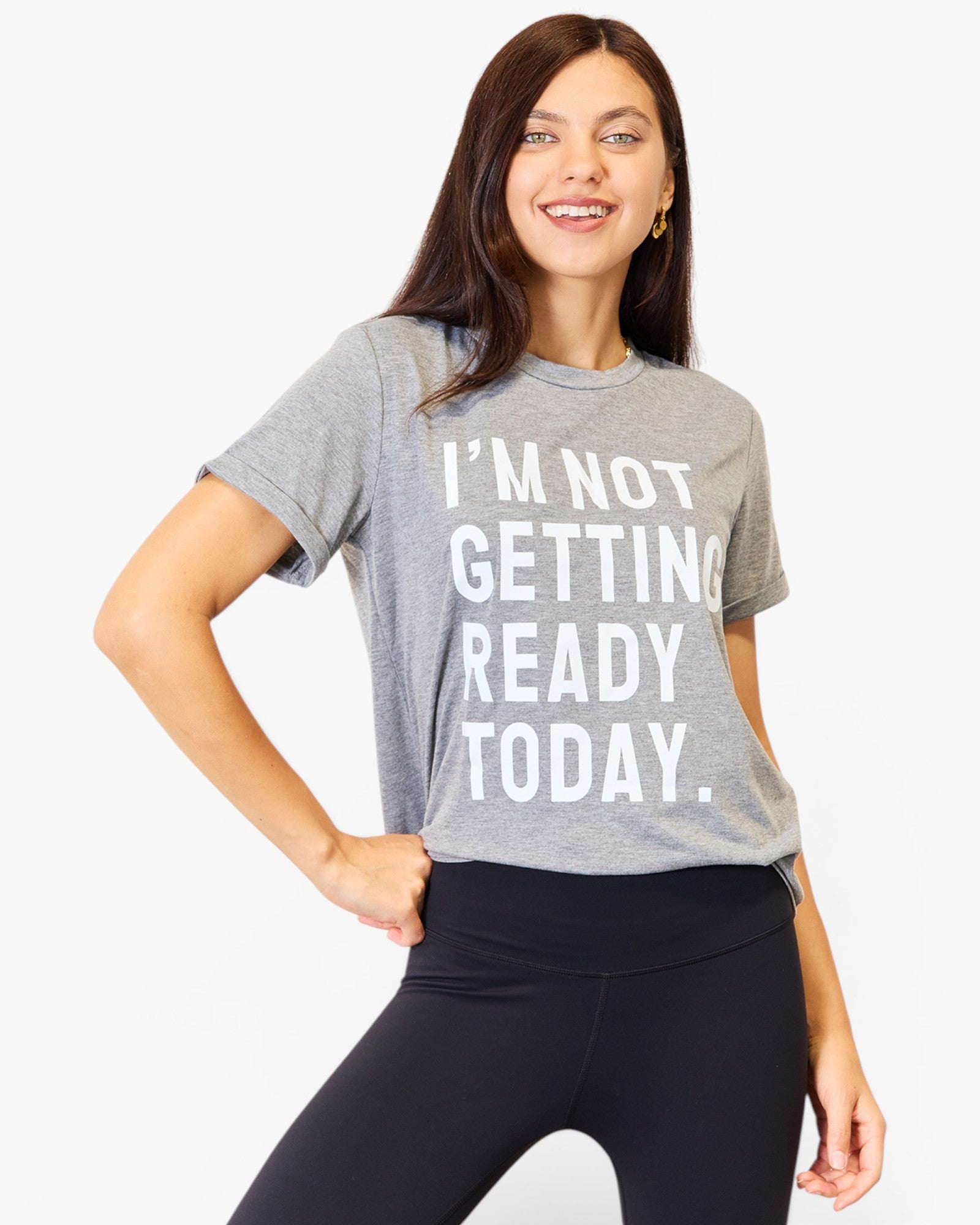 A woman wearing a heather gray graphic t-shirt with the text 'I'M NOT GETTING READY TODAY.' in bold white letters, paired with black leggings, against a plain white background.
