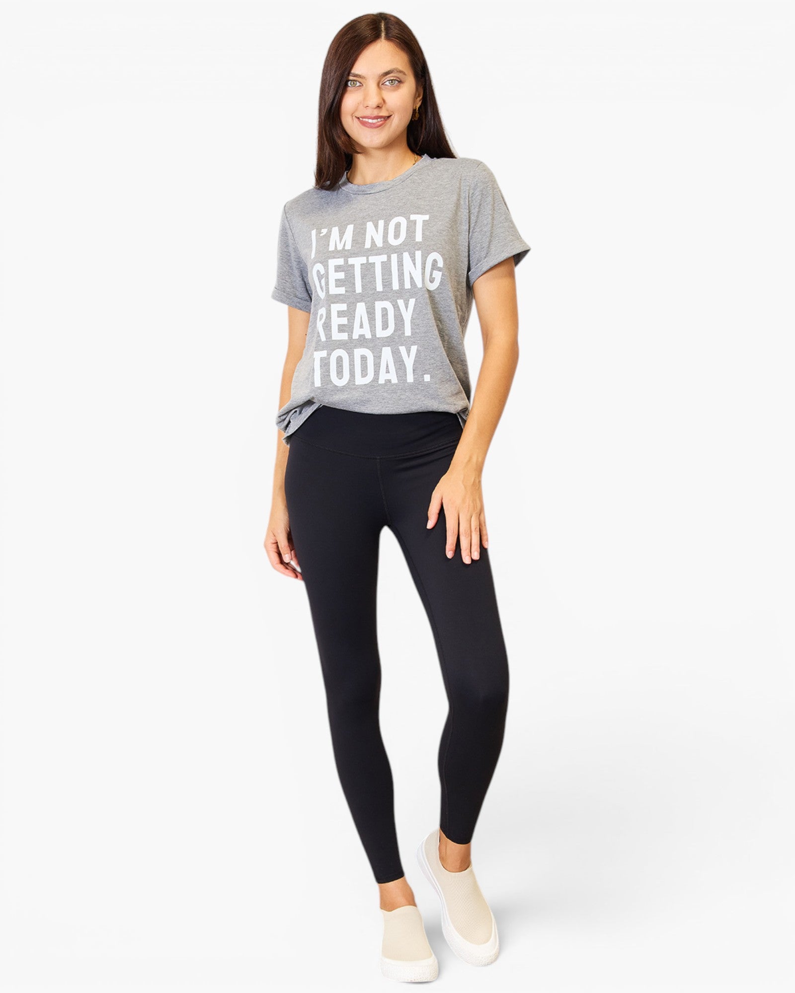 A woman modeling a heather gray graphic t-shirt with white text that reads 'I'M NOT GETTING READY TODAY.' paired with black leggings and white slip-on sneakers, against a plain white studio background.