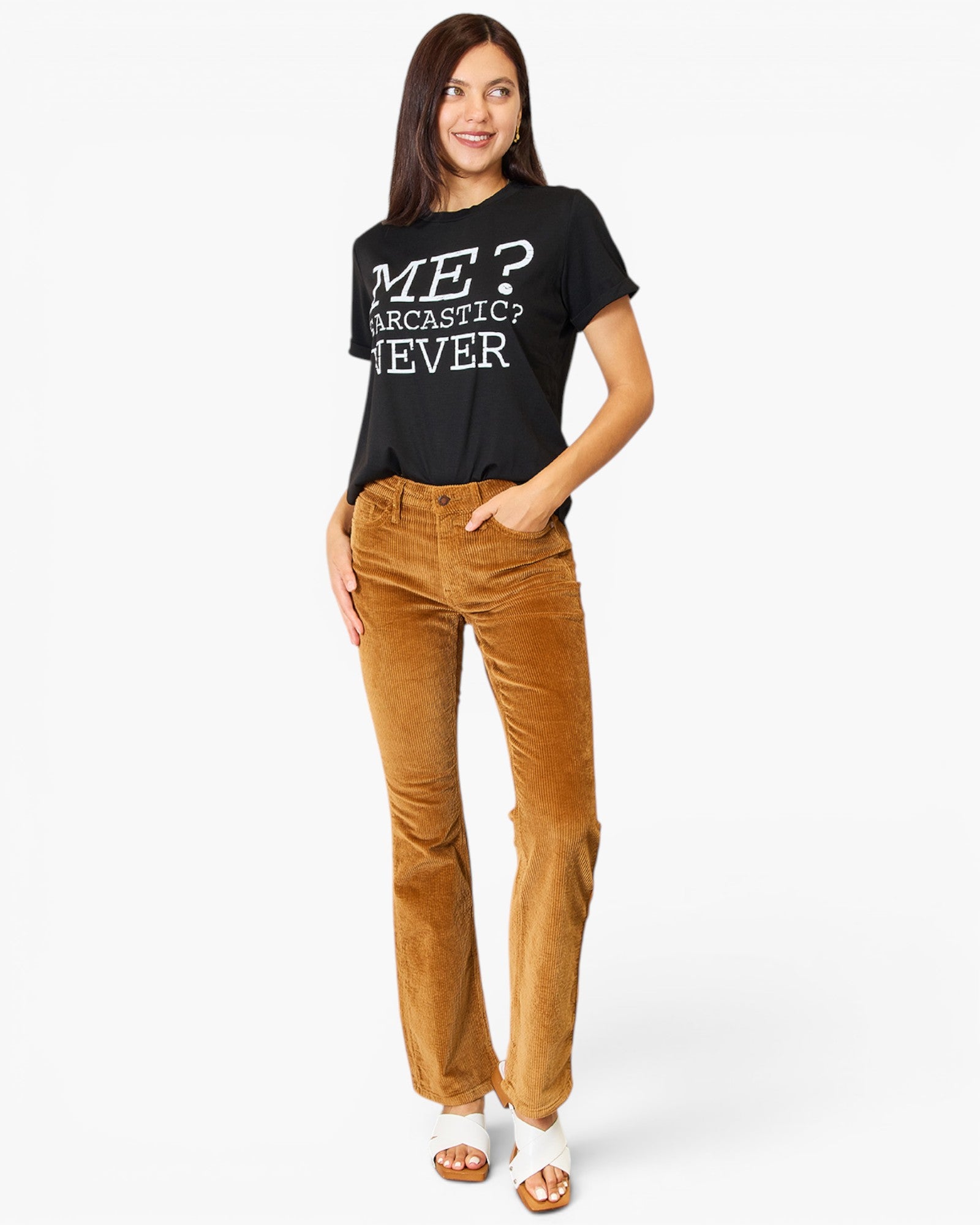 A woman modeling a black graphic t-shirt with white text that reads 'ME? SARCASTIC? NEVER' paired with brown corduroy pants and white sandals, set against a plain white studio background.