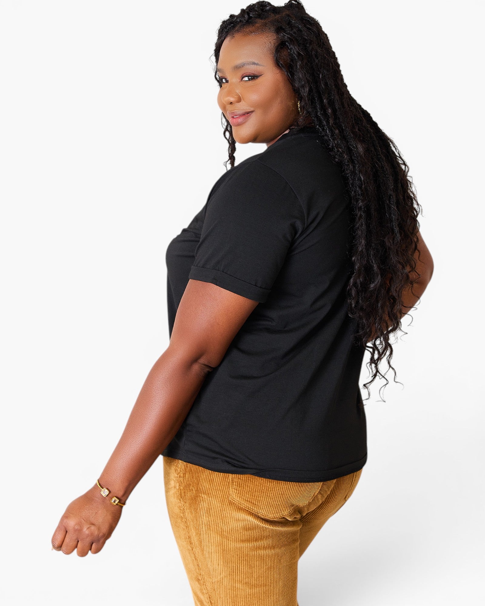 A woman wearing a black short-sleeve t-shirt and mustard yellow corduroy pants, shown from a side-back angle against a light gray studio background.