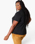 A woman wearing a black short-sleeve t-shirt and mustard yellow corduroy pants, shown from a side-back angle against a light gray studio background.