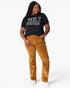 A woman modeling a black graphic t-shirt with white text that reads 'ME? SARCASTIC? NEVER' and mustard brown corduroy pants, standing against a light gray studio background.