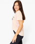 A woman with long brown hair is shown from the side/back wearing a light peach-pink short-sleeve crop top with a black graphic print and black leggings, set against a plain white studio background.