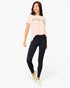 A woman modeling a light pink short-sleeve t-shirt with black script lettering on the chest, paired with black leggings and beige slip-on shoes, against a plain white background.