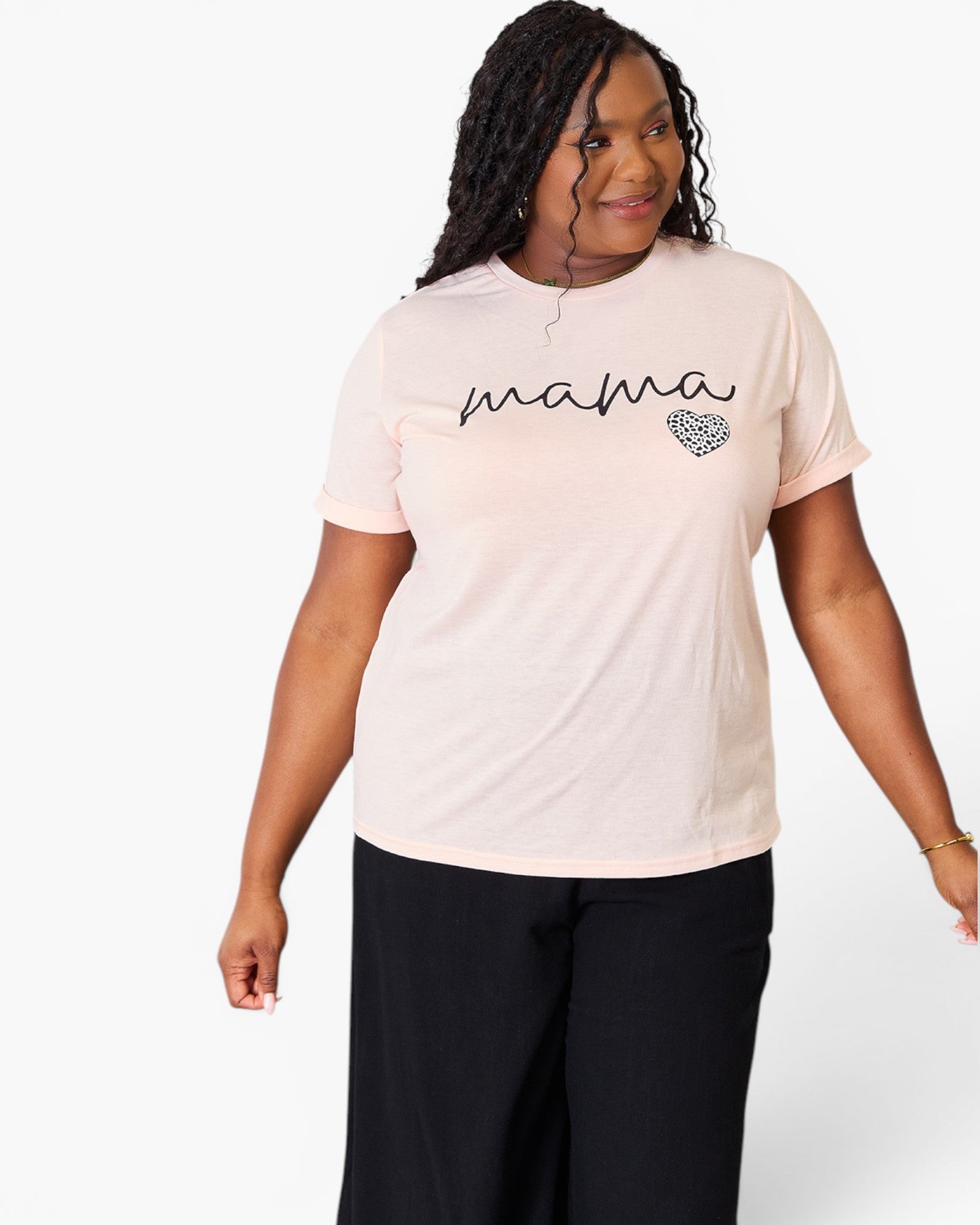 A woman modeling a light pink short-sleeve t-shirt with 'mama' written in black script and a small heart graphic on the chest, paired with black pants against a plain white background.