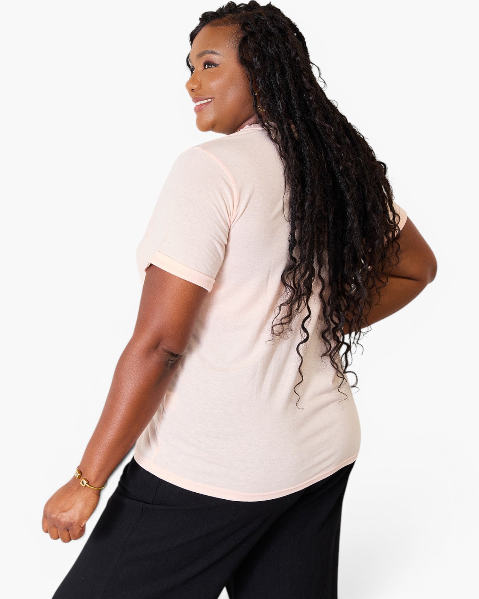 A woman with long, curly black hair is shown from a back-three-quarter angle wearing a light peach-pink short-sleeve crewneck t-shirt and black pants against a plain white studio background.