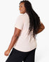 A woman with long, curly black hair is shown from a back-three-quarter angle wearing a light peach-pink short-sleeve crewneck t-shirt and black pants against a plain white studio background.