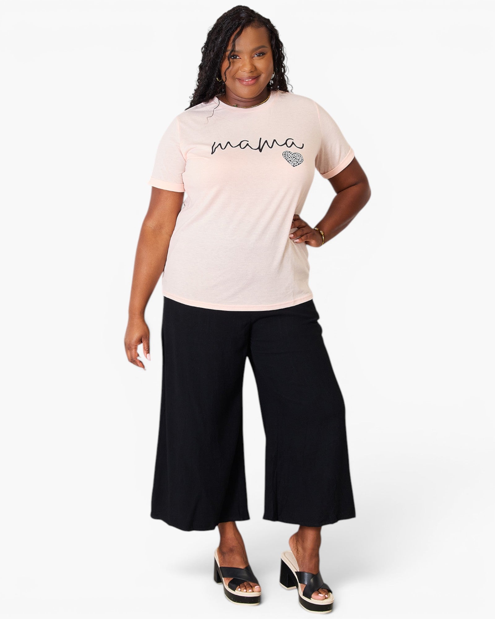 A woman modeling a light pink short-sleeve t-shirt with 'mama' and a small heart graphic printed on the chest, paired with black wide-leg cropped pants and black heeled sandals, against a plain white studio background.