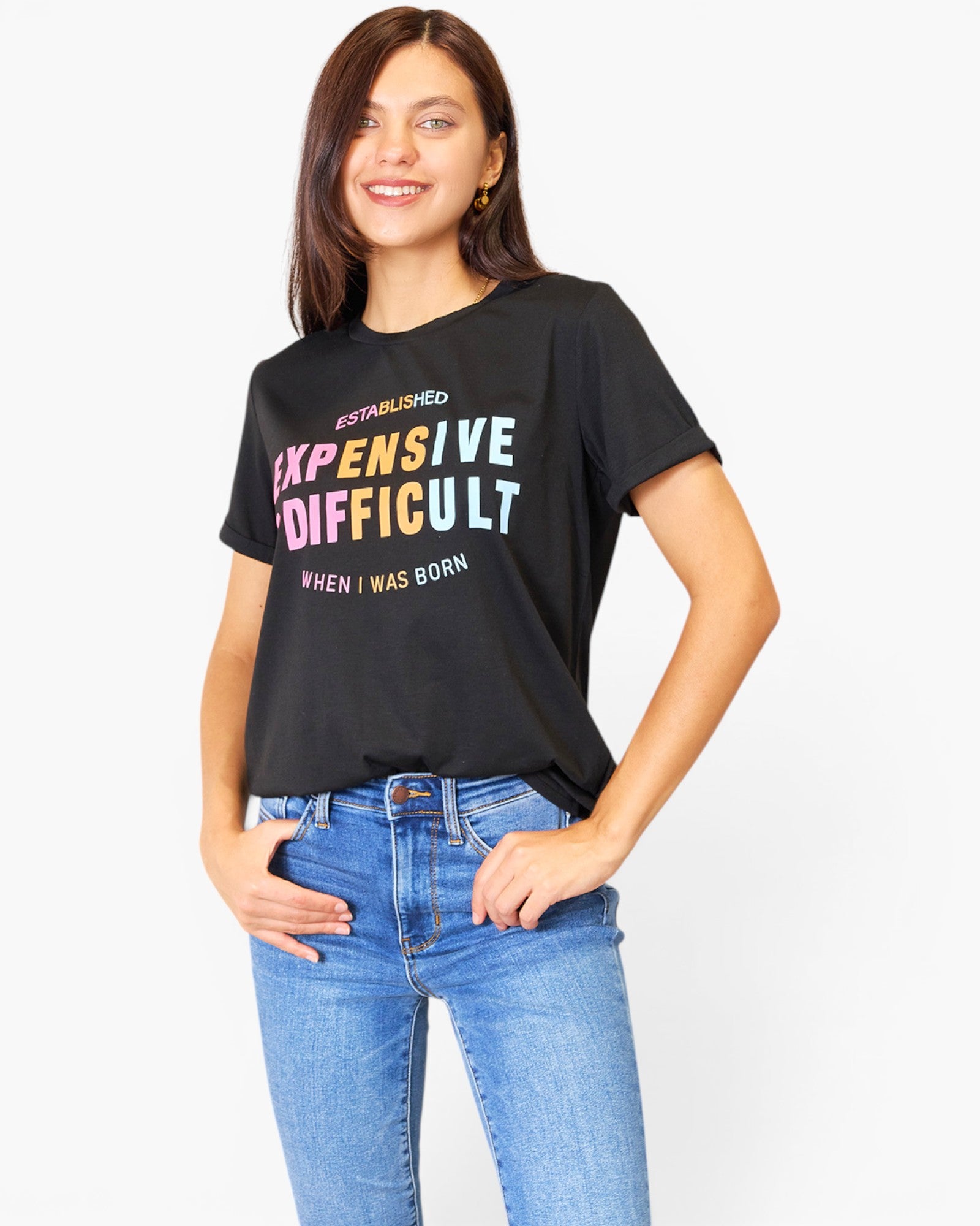 A woman wearing a black graphic t-shirt with the text 'EXPENSIVE DIFFICULT' in a rainbow gradient and 'ESTABLISHED WHEN I WAS BORN' in white, paired with blue jeans, against a plain white background.