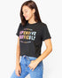 A woman wearing a black graphic t-shirt with colorful text that reads 'EXPENSIVE DIFFICULT WHEN I WAS BORN' and blue jeans, posing against a light gray background.