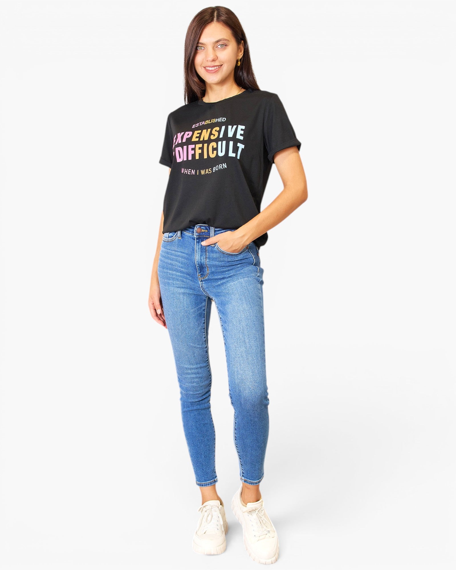A woman modeling a black graphic t-shirt with the text 'EXPENSIVE DIFFICULT' in white, pink, and yellow, paired with blue skinny jeans and white sneakers, against a plain white background.