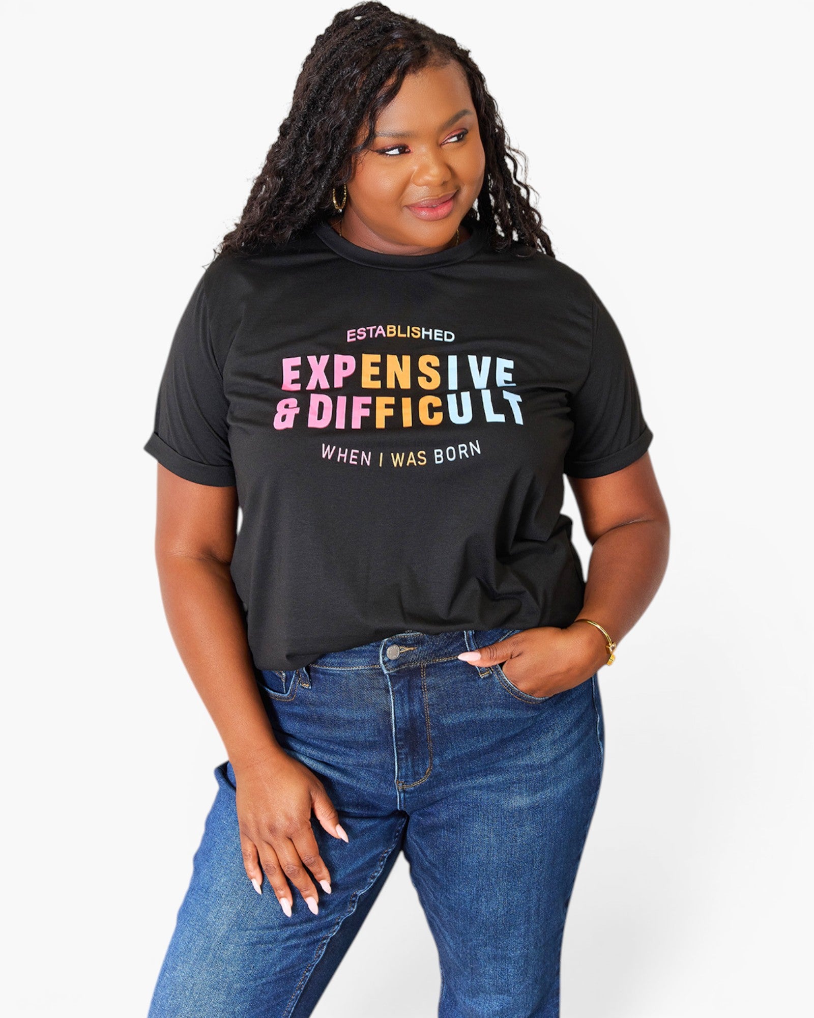 A woman wearing a black graphic t-shirt with the text 'ESTABLISHED EXPENSIVE & DIFFICULTY WHEN I WAS BORN' in pink, blue, and white, paired with blue jeans, against a plain white background.