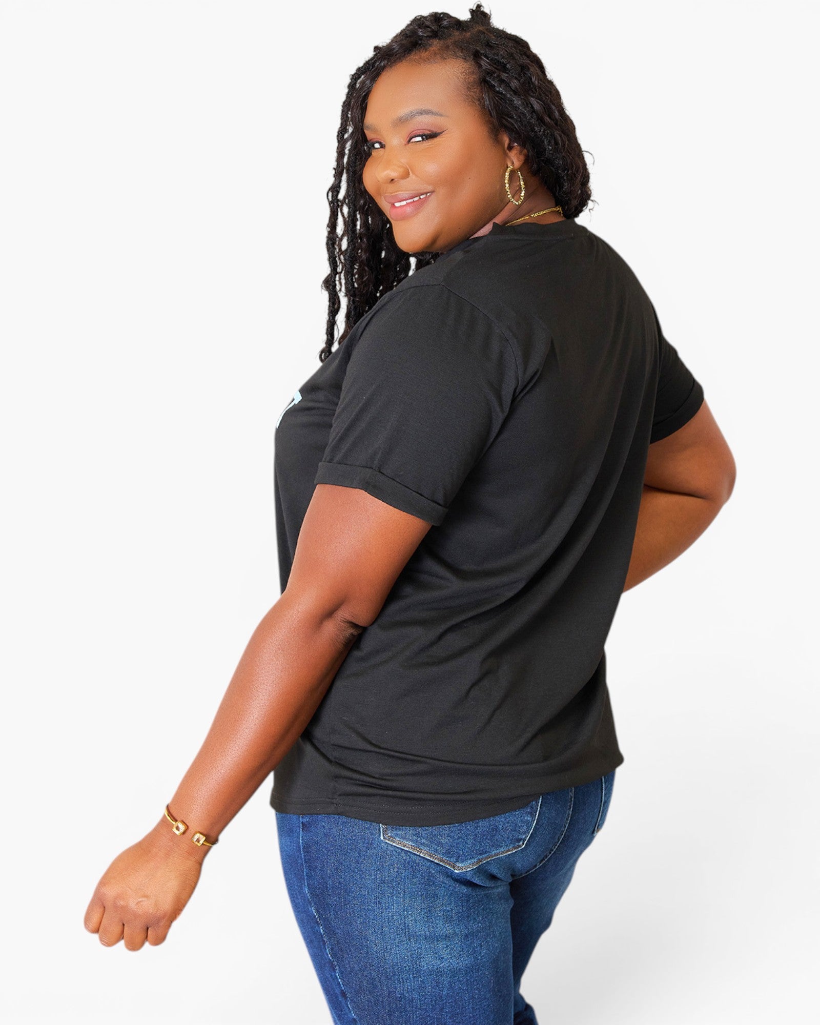 A woman with long black braids is shown from a three-quarters back view, wearing a black short-sleeve t-shirt and blue jeans against a plain white studio background.