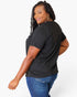 A woman with long black braids is shown from a three-quarters back view, wearing a black short-sleeve t-shirt and blue jeans against a plain white studio background.
