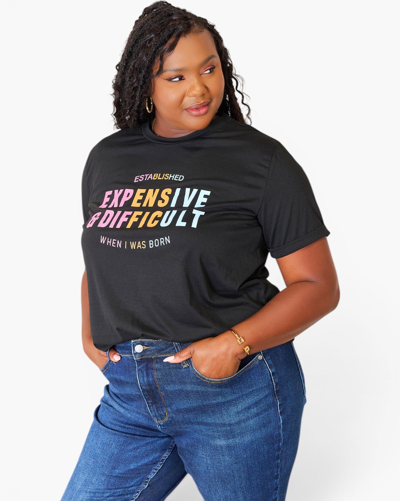 A woman wearing a black graphic t-shirt with the text 'EXPENSIVE & DIFFICULT WHEN I WAS BORN' in white, yellow, and pink lettering, paired with blue jeans, against a plain white background.