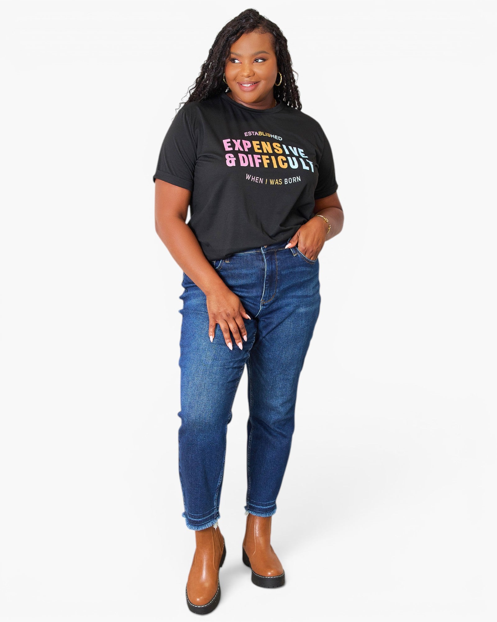 A woman modeling a black graphic t-shirt with colorful text paired with blue jeans and brown ankle boots, against a plain white background.