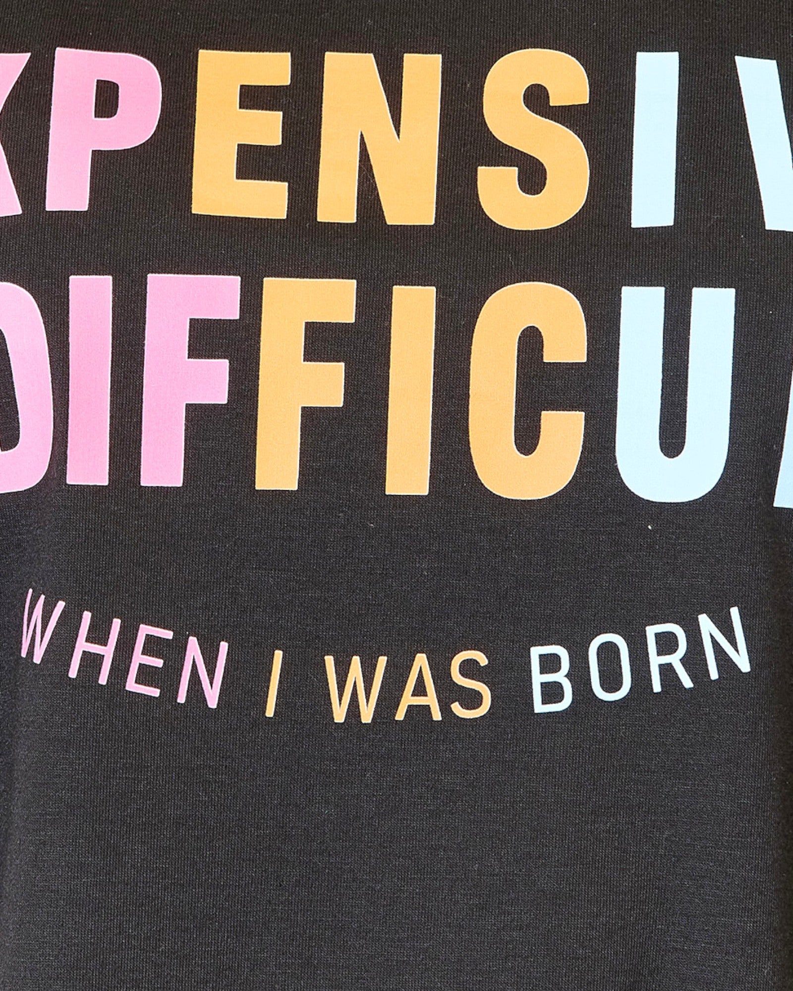 Close-up of a black graphic T-shirt with large pastel text reading 'EXPENSIVE & DIFFICULT EST  WHEN I WAS BORN' in pink, yellow, and light blue lettering.