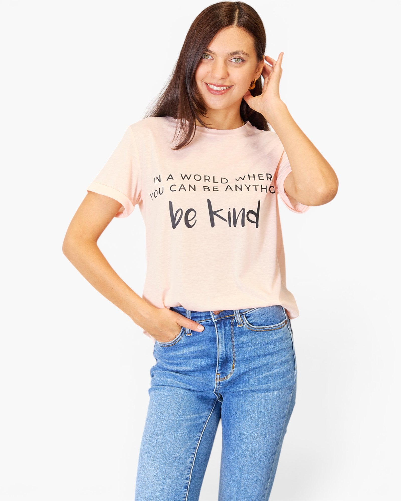 A woman modeling a light pink graphic t-shirt with the text 'IN A WORLD WHERE YOU CAN BE ANYTHING be kind' in black lettering, paired with blue jeans against a plain white background.