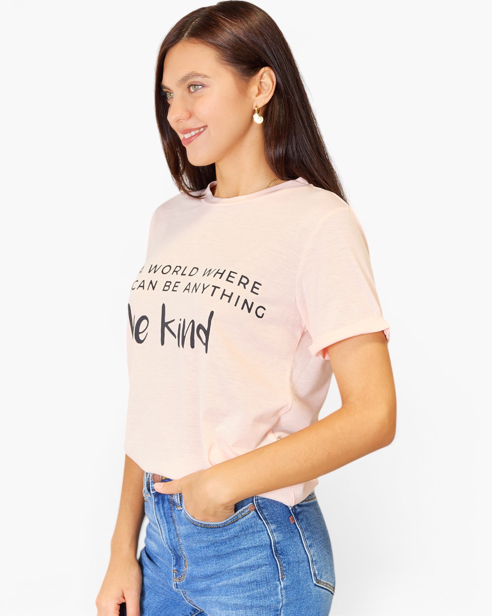 A woman wearing a light pink short-sleeve t-shirt with black text that reads 'IN A WORLD WHERE ANYONE CAN BE ANYTHING we kind' paired with blue jeans, set against a plain white background.