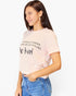 A woman wearing a light pink short-sleeve t-shirt with black text that reads 'IN A WORLD WHERE ANYONE CAN BE ANYTHING we kind' paired with blue jeans, set against a plain white background.