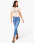 A woman modeling a light pink graphic t-shirt with the text 'A WORLD WHERE I CAN BE ANTI' and 'be kind' printed in black, paired with blue jeans and brown ankle boots, against a plain white background.