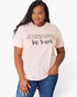 A woman modeling a light pink graphic t-shirt with the text 'IN A WORLD WHERE YOU CAN BE ANYTHING. be kind' printed in black, paired with blue jeans against a plain white background.