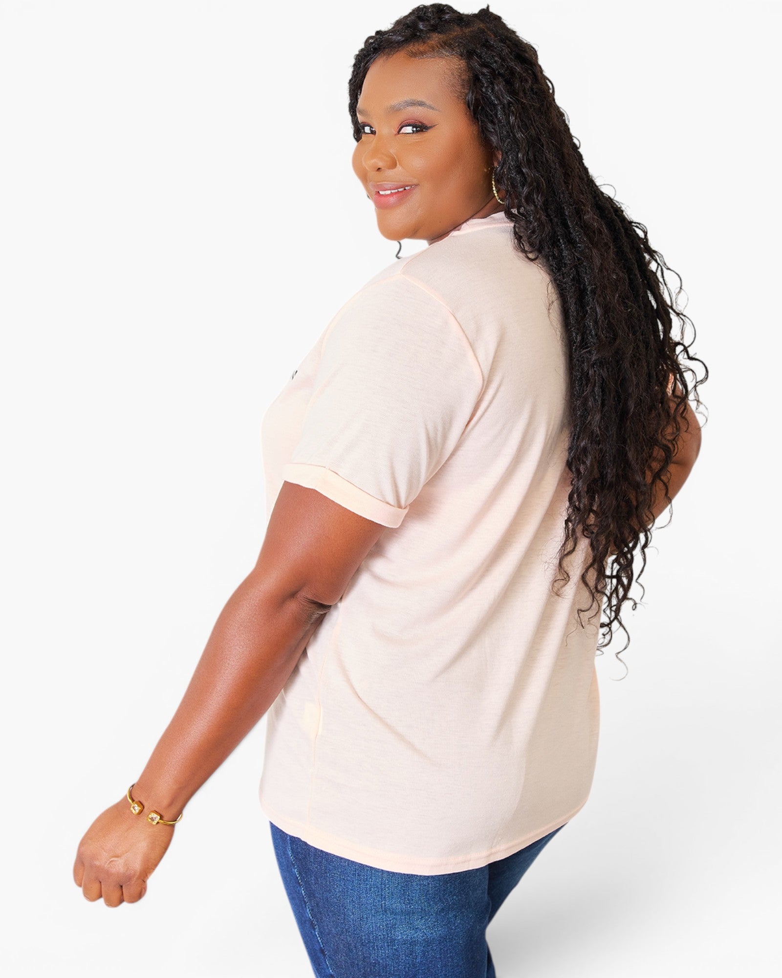 A woman with long, curly black hair is shown from a side-back angle wearing a light peach-pink short-sleeve t-shirt and blue jeans against a plain white studio background.