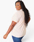A woman with long, curly black hair is shown from a side-back angle wearing a light peach-pink short-sleeve t-shirt and blue jeans against a plain white studio background.