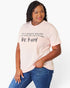 A woman modeling a light pink graphic t-shirt with the text 'IN A WORLD WHERE YOU CAN BE ANYTHING. be kind' printed in black, paired with blue jeans against a plain white background.