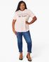 A woman modeling a light pink graphic t-shirt with the text 'be kind' and 'IN A WORLD WHERE EVERYONE IS TRYING TO BE KIND' printed on the chest, paired with blue jeans and brown ankle boots against a white studio background.