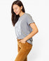 A woman wearing a heather gray t-shirt with a white distressed American flag graphic and brown corduroy pants, posing against a light gray studio background.